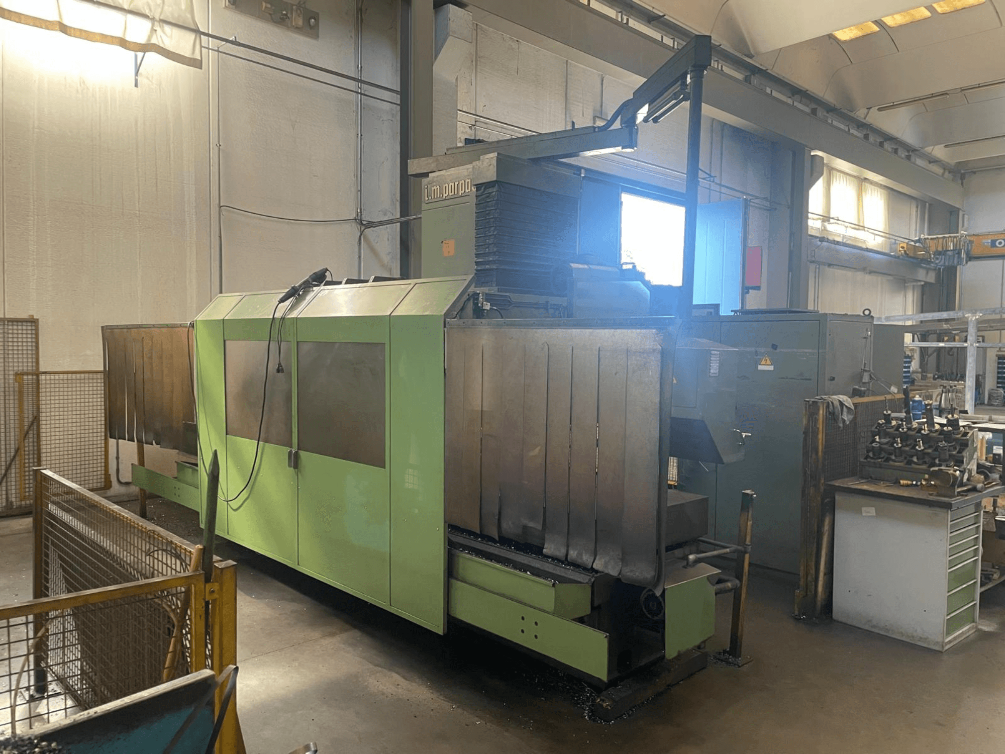 Зеленый промышленный станок с видимыми металлическими компонентами, вид спереди; панель управления, предупреждения о безопасности и рабочее место рядом.