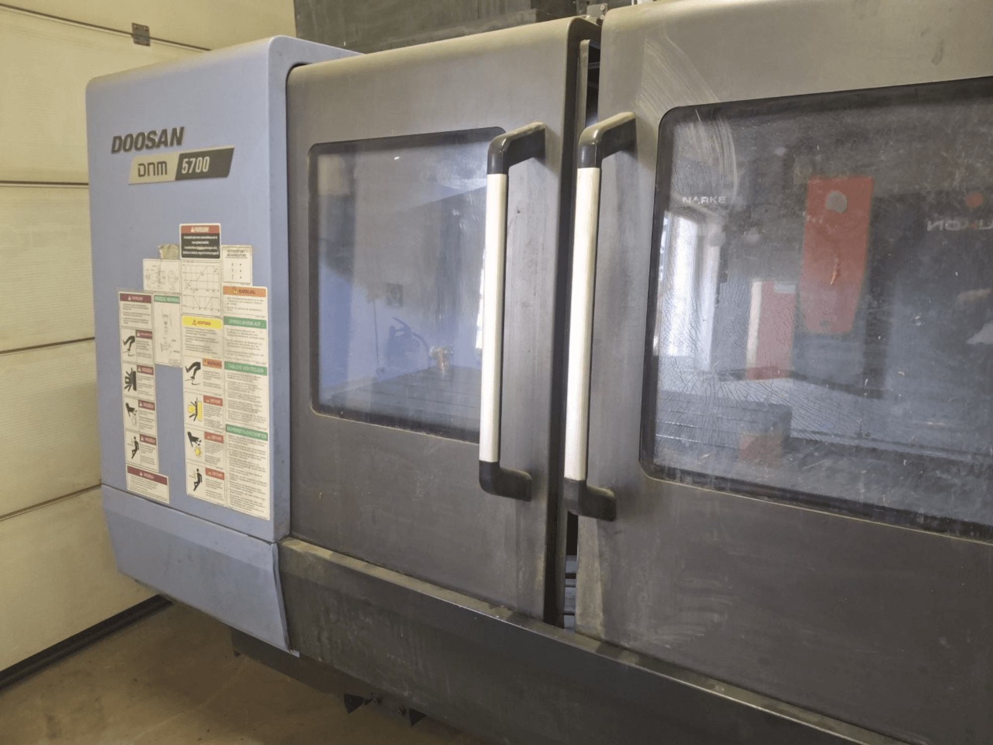 Вид спереди на вертикальный обрабатывающий центр DOOSAN DNM 5700 с двойными дверями и панелями управления с инструкциями по эксплуатации.