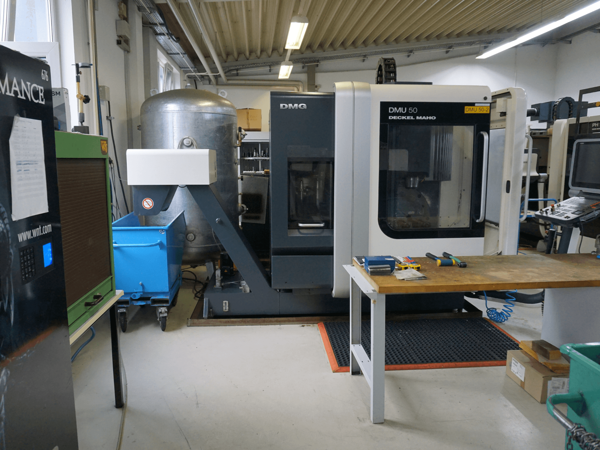 Фрезерный станок DMG DMU 50 в мастерской, вид спереди, с инструментами на деревянном столе и синей корзиной на колесиках рядом.