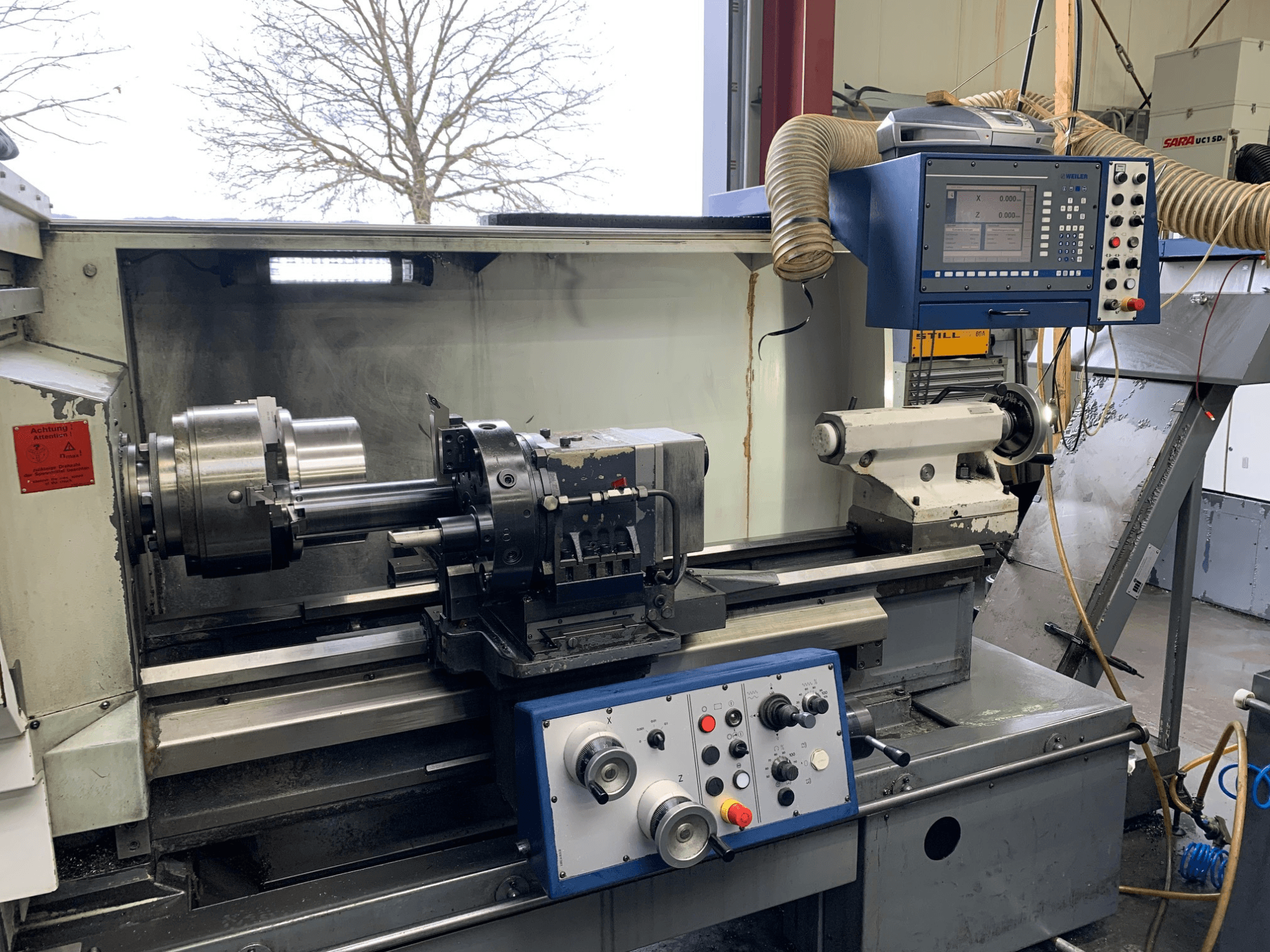 Горизонтальный токарный станок Weiler E55X1000 в мастерской, на котором видны панель управления, токарный станок и держатель инструмента. Вид спереди.