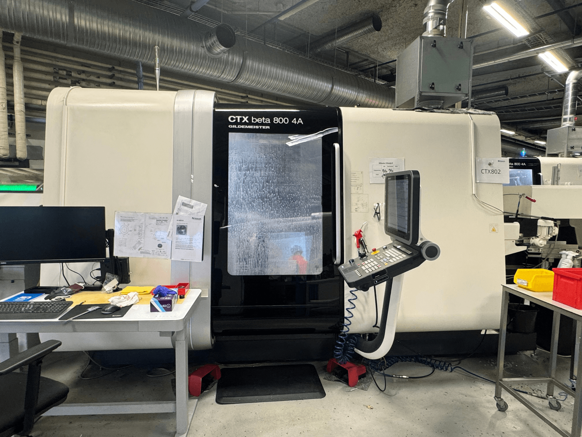 Станок с ЧПУ CTX beta 800 4A в мастерской, вид сбоку на панель управления и рабочую зону, загроможденную инструментами и документами.
