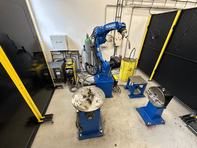 Роботизированный манипулятор Yaskawa синего цвета с двумя рабочими столами, сварочным оборудованием и защитными ограждениями; вид спереди на промышленное рабочее место.