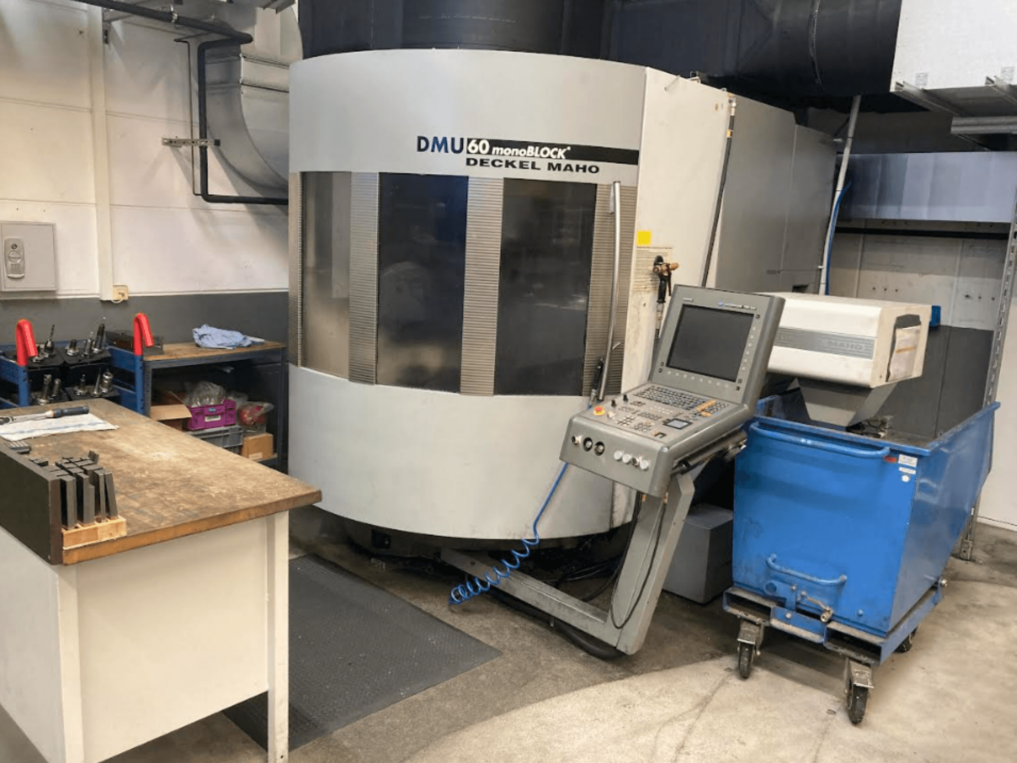 Моноблочный вертикальный обрабатывающий центр DMG MORI DMU 60, вид спереди с панелью управления, рабочей зоной и синей тележкой для инструментов.