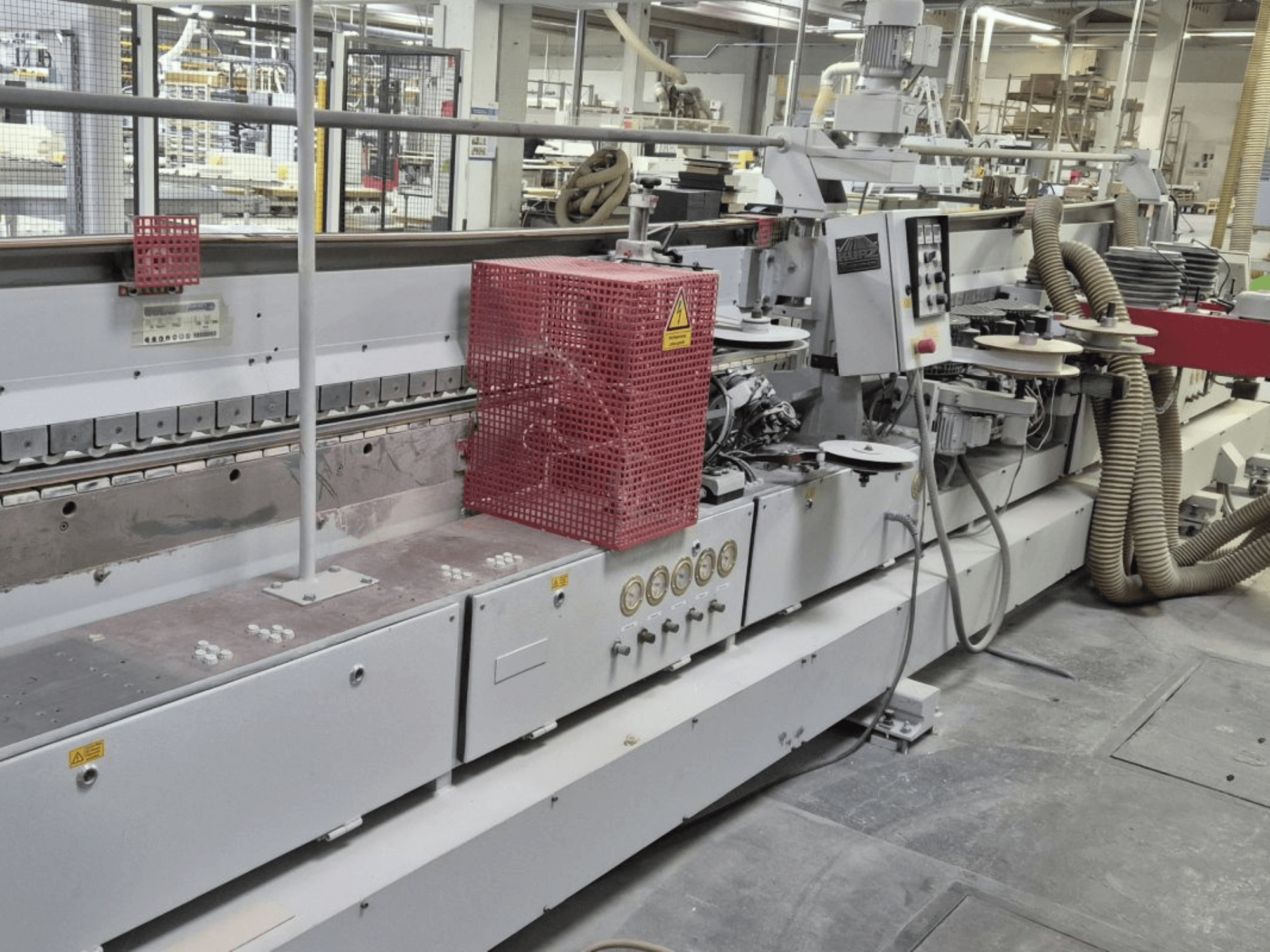 Кромкооблицовочный станок с панелью управления, всасывающими шлангами и красным защитным кожухом, вид сбоку.