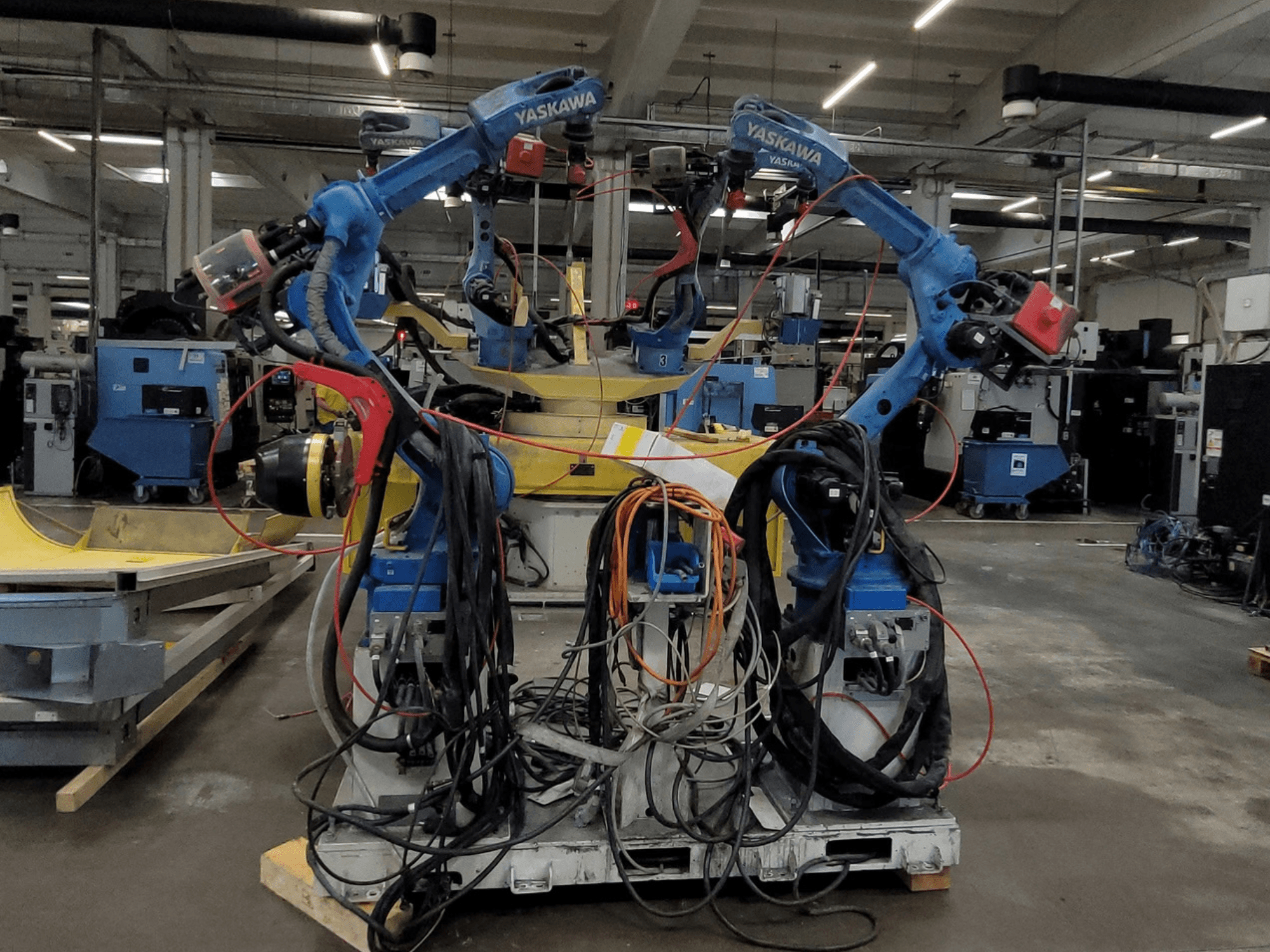 Роботизированная сварочная ячейка Yaskawa RWC L1_OS 15330, вид спереди, с двумя синими роботами, оснащёнными длинными кабелями и сварочным оборудованием.