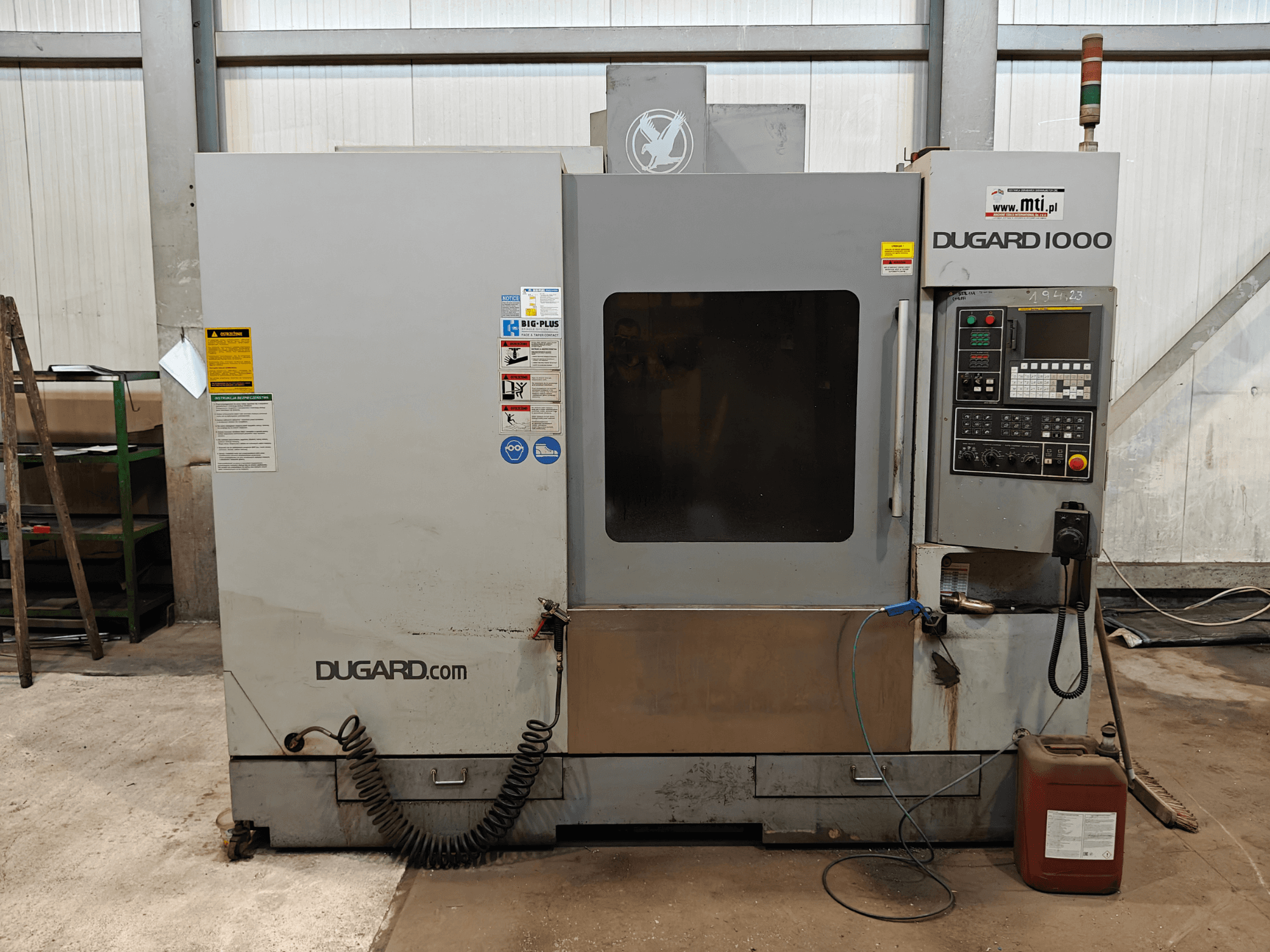 Станок с ЧПУ Dugard 1000, вид спереди, на панели управления, наклейки безопасности и контейнер для масла, в мастерской.
