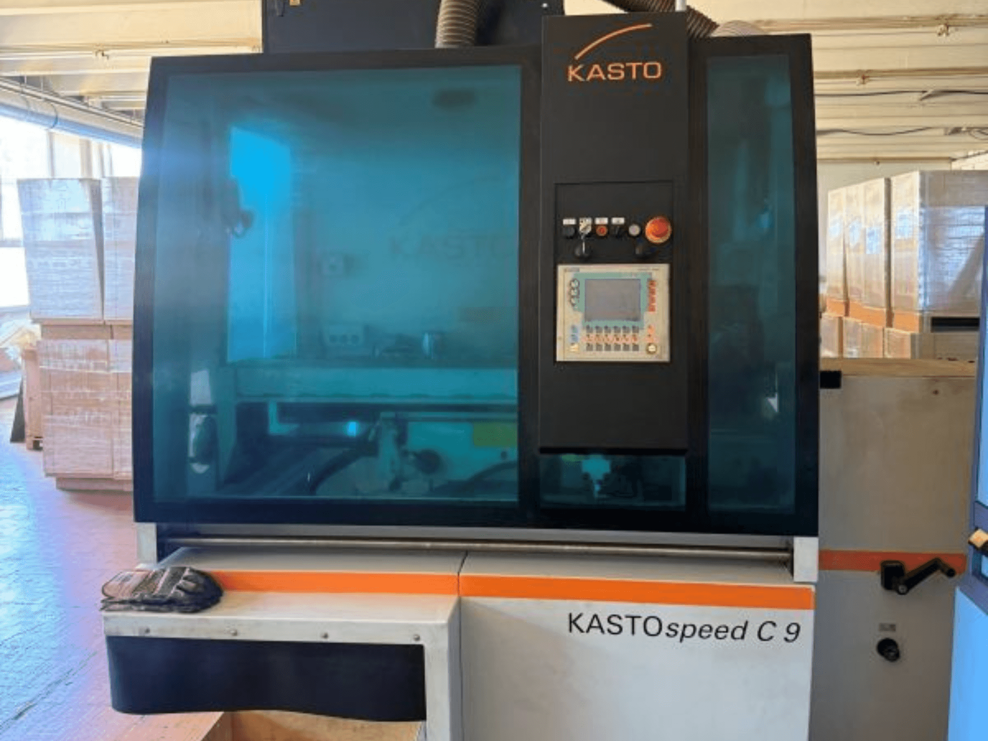 Вид спереди на циркулярную пилу для резки металла KASTO KASTOspeed C 9 с панелью управления и защитным стеклом; на заднем плане видны деревянные ящики.