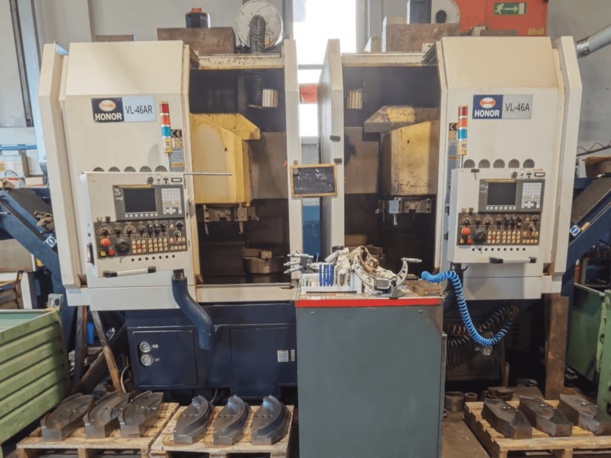 Вид спереди вертикального токарного станка HONOR SEIKI VL 46A с панелью управления и сопутствующими деталями, размещенными на деревянных поддонах.