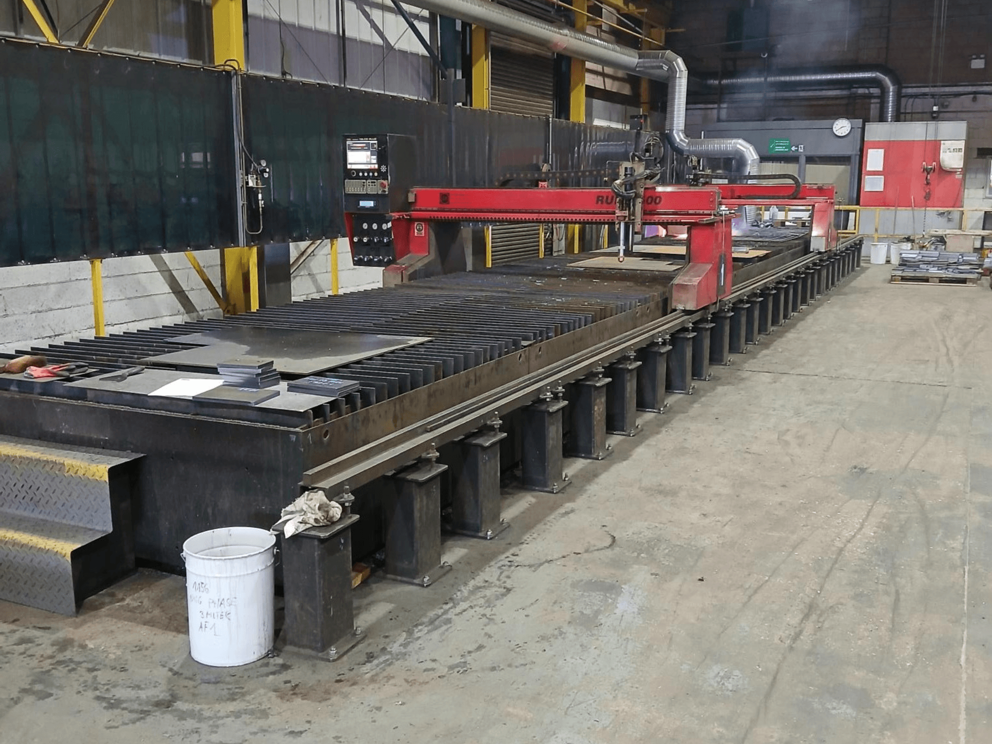 Красный станок для резки RUIDA 300, вид сбоку, с металлическим листом на стальном столе, расположенный в промышленном рабочем пространстве.