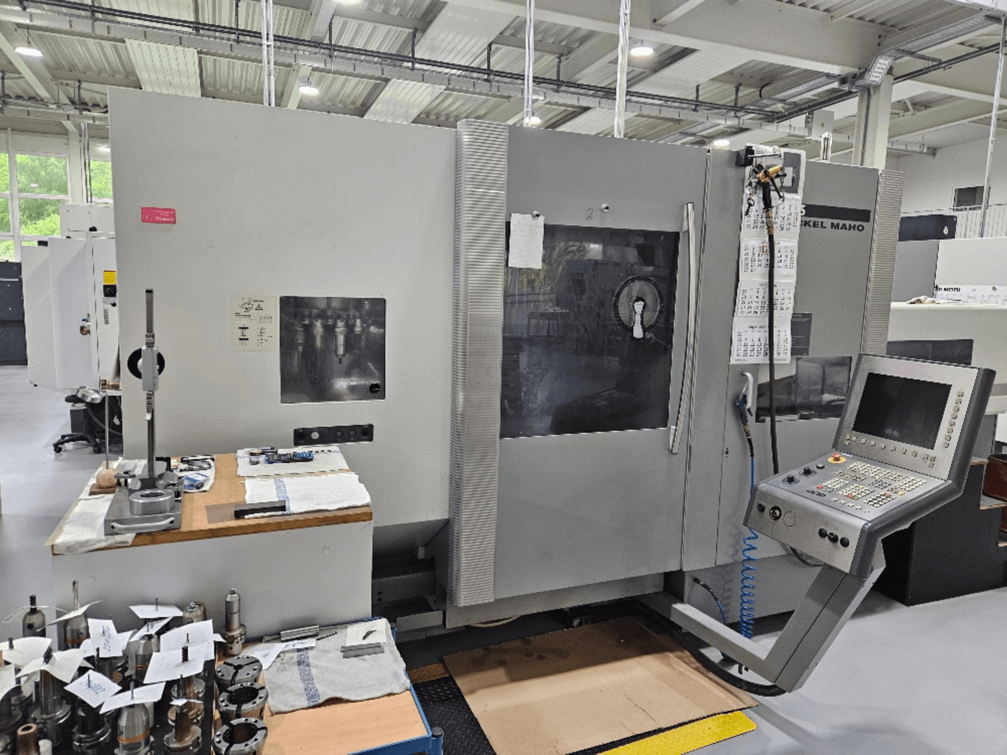 Вид спереди на станок DMG Mori DMU 70 с панелью управления, инструментами на деревянном столе и различными заготовками рядом.