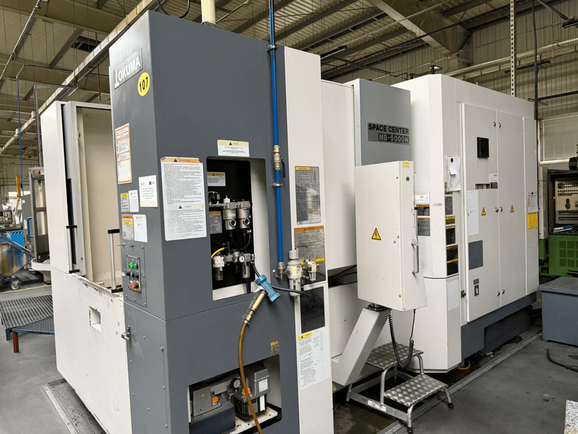 Горизонтальный обрабатывающий центр Okuma MB 5000H, вид сбоку, демонстрирующий панель управления и компоненты оборудования в заводских условиях.