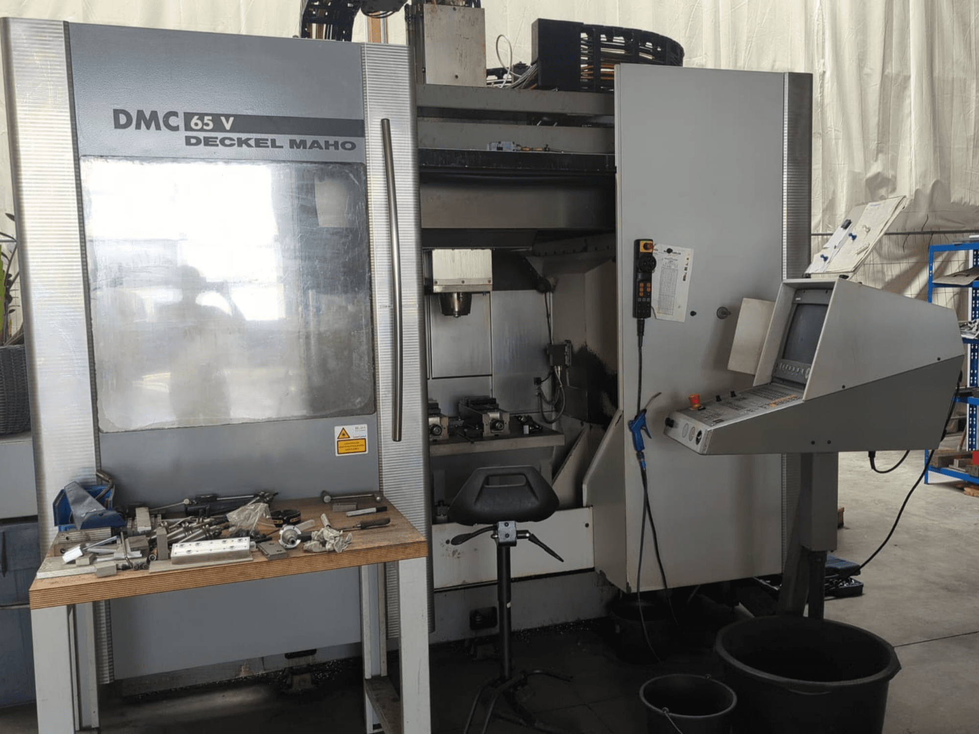 Вид спереди на вертикальный обрабатывающий центр DECKEL MAHO DMC 65 V с панелью управления, инструментами на столе и подготовленной рабочей зоной.