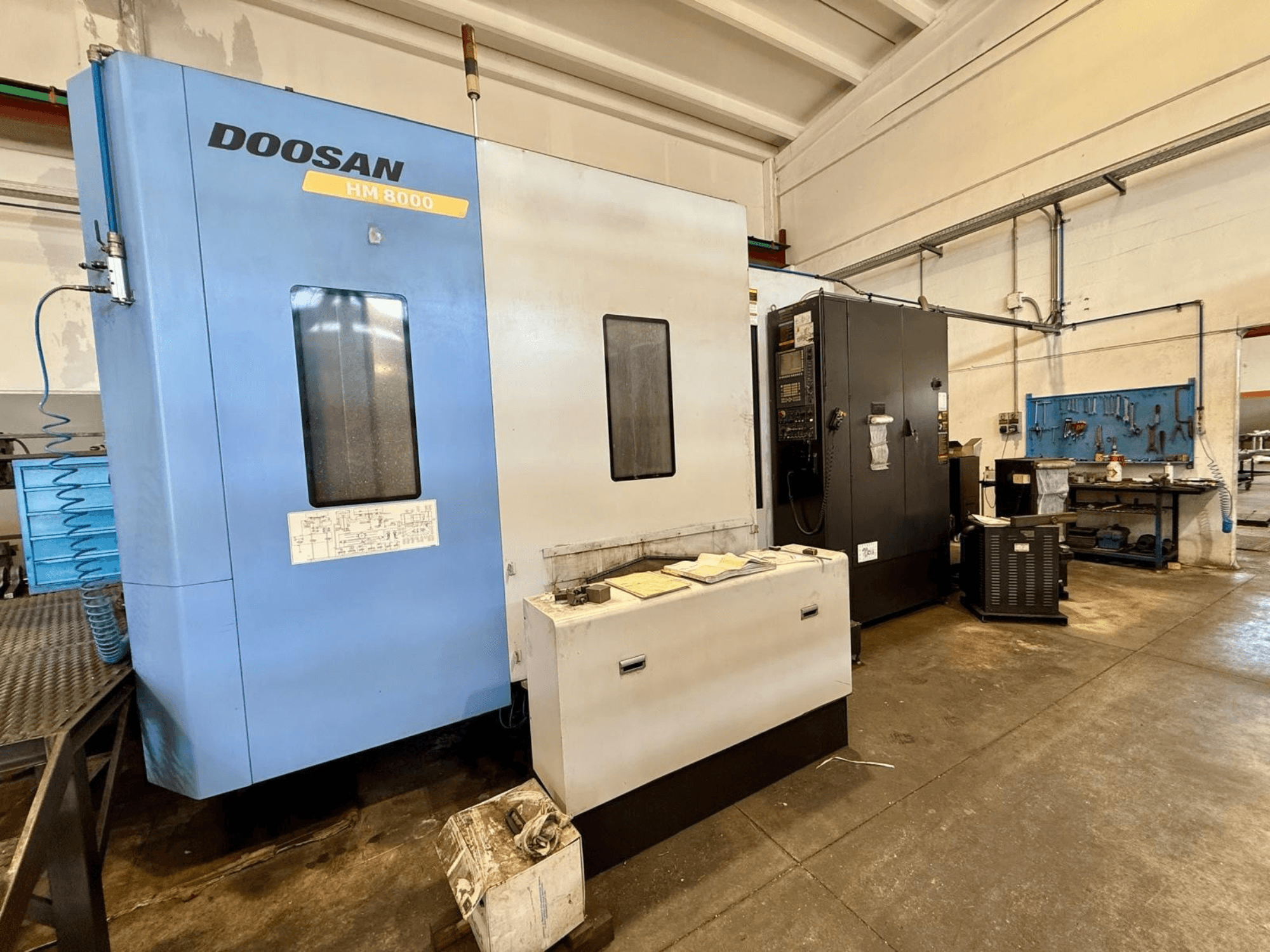 Горизонтальный обрабатывающий центр DOOSAN HM 8000 синего цвета, вид спереди, на котором видны панель управления и расположенное рядом цеховое оборудование.