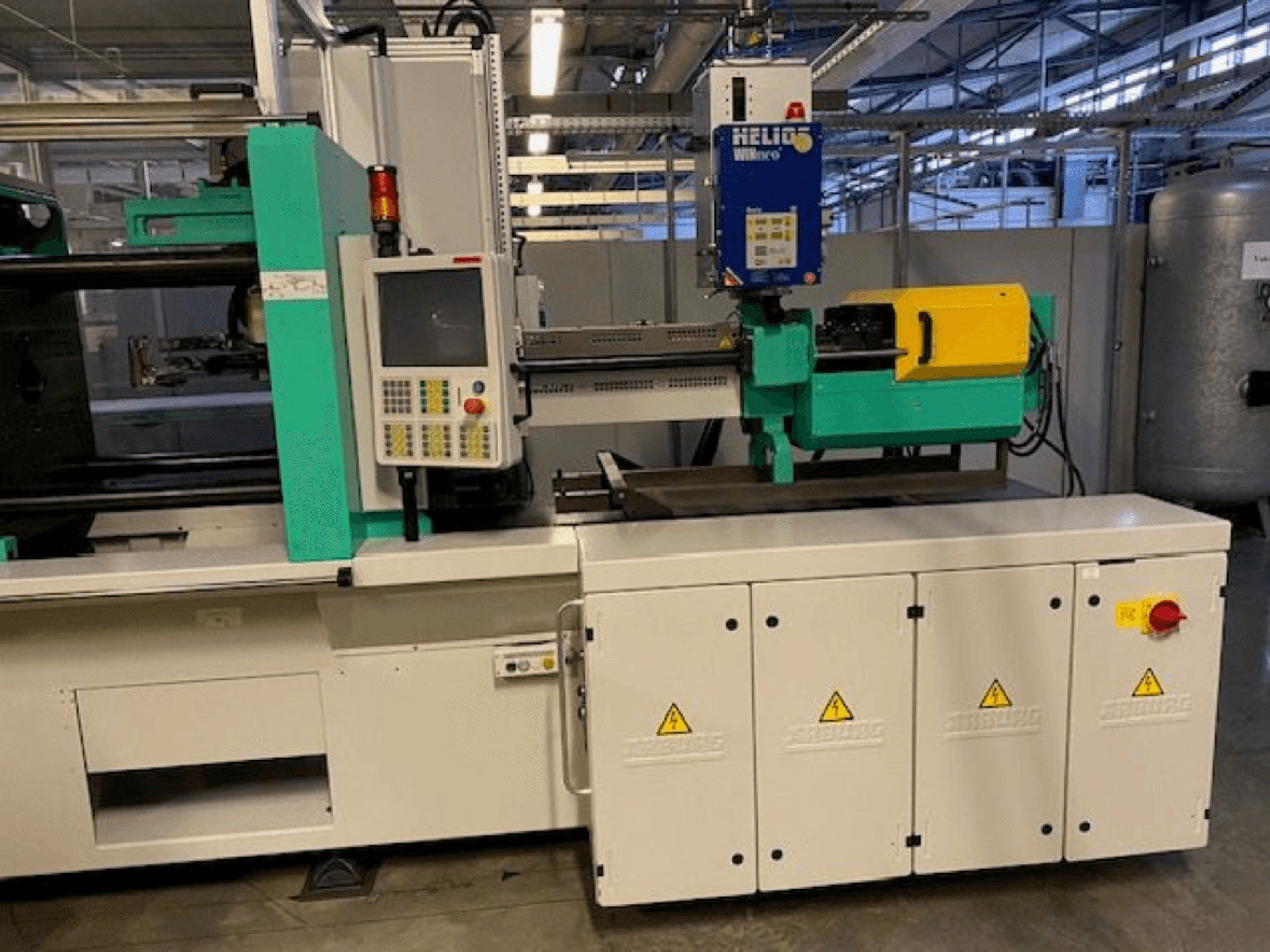 Промышленная машина для литья под давлением, модель HELIO, вид спереди, панель управления, защитные ограждения и кнопка аварийной остановки.