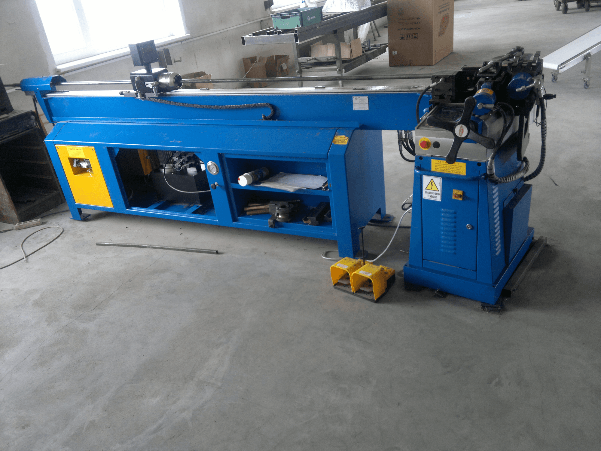 Синий деревообрабатывающий станок с желтыми акцентами, вид сбоку; имеет панель управления, ножные педали и вращающийся узел ножа.