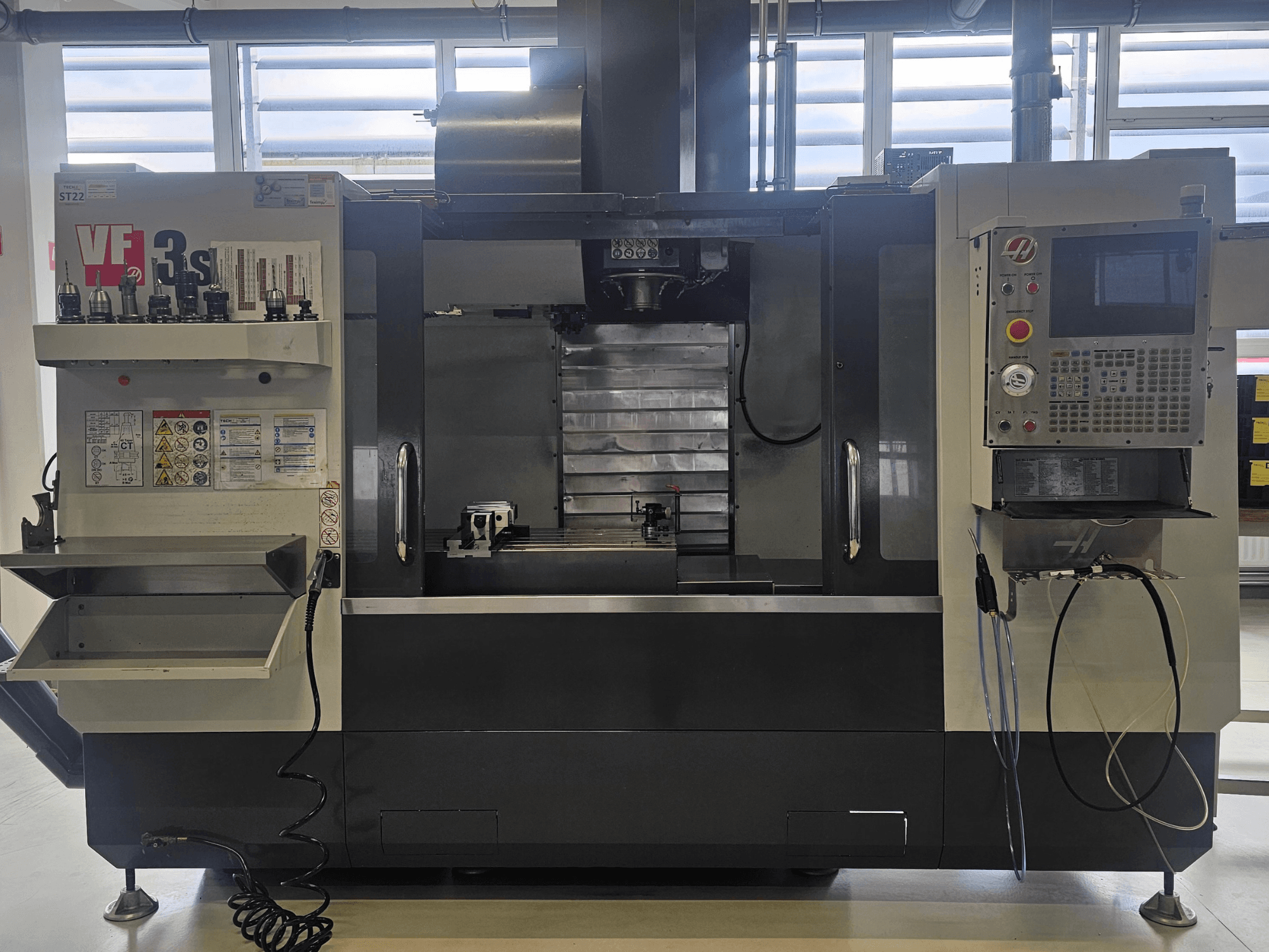 Фрезерный станок с ЧПУ Haas VF3SS, вид спереди, видны держатели инструментов, панель управления и пустой поддон для охлаждающей жидкости, видны наклейки безопасности.