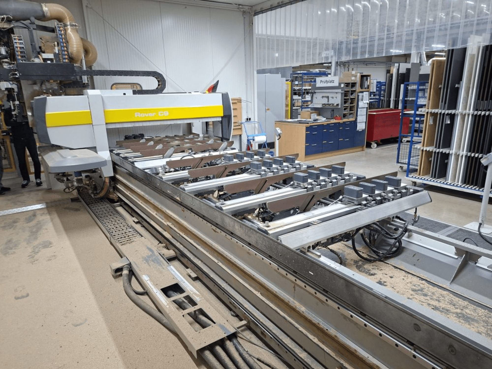 Станок с ЧПУ Rover C9 в мастерской; вид спереди показывает режущие инструменты и конвейерную систему, на полу скапливается пыль.