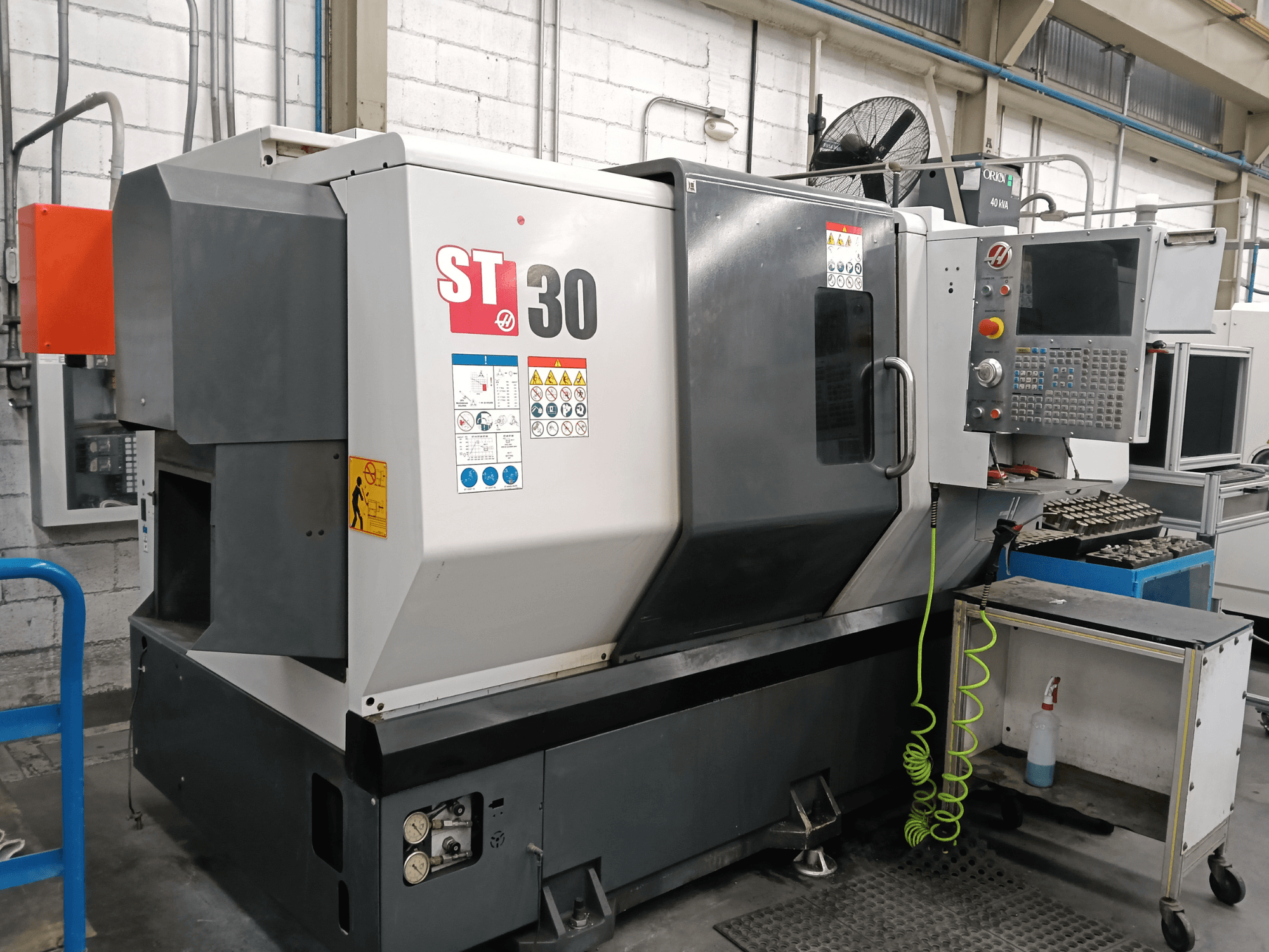Альтернативный текст: Вид спереди на горизонтальный токарный станок HAAS ST-30 с панелью управления, предупреждающими надписями и инструментами на стоящей рядом тележке.