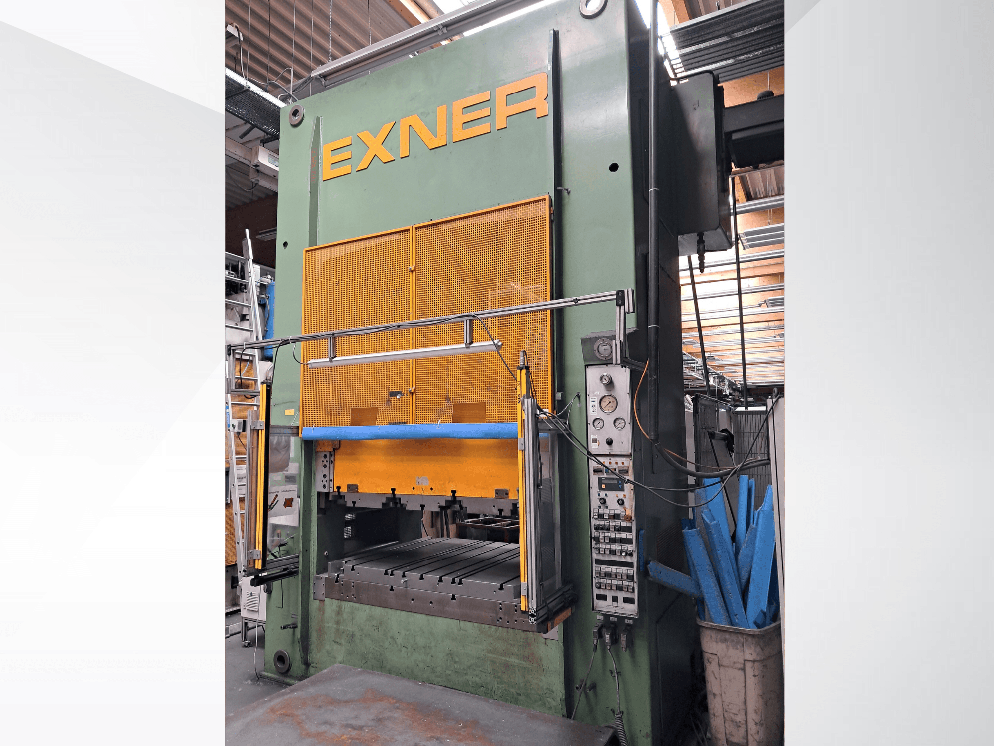 Гидравлический пресс EXNER EX SBR 150, вид спереди; с органами управления, синей защитной перекладиной и сетчатой защитной решеткой, установленный в цехе.