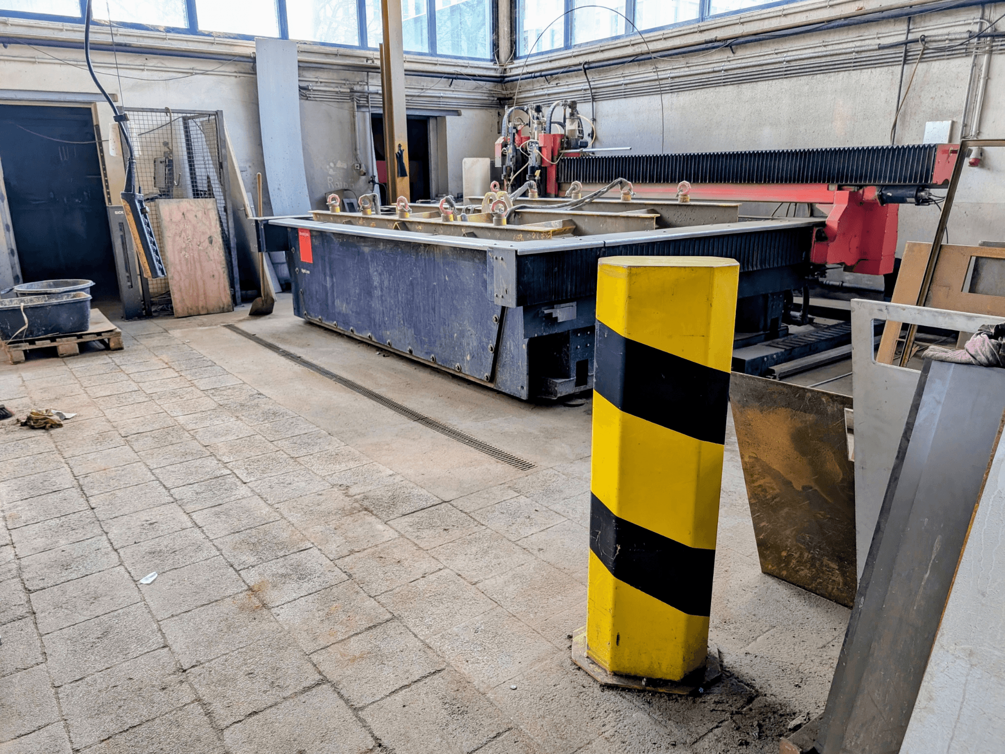 Станок гидроабразивной резки Bystronic Byjet L 2030 в мастерской, вид сбоку, с желтым столбом безопасности и окружающими материалами.