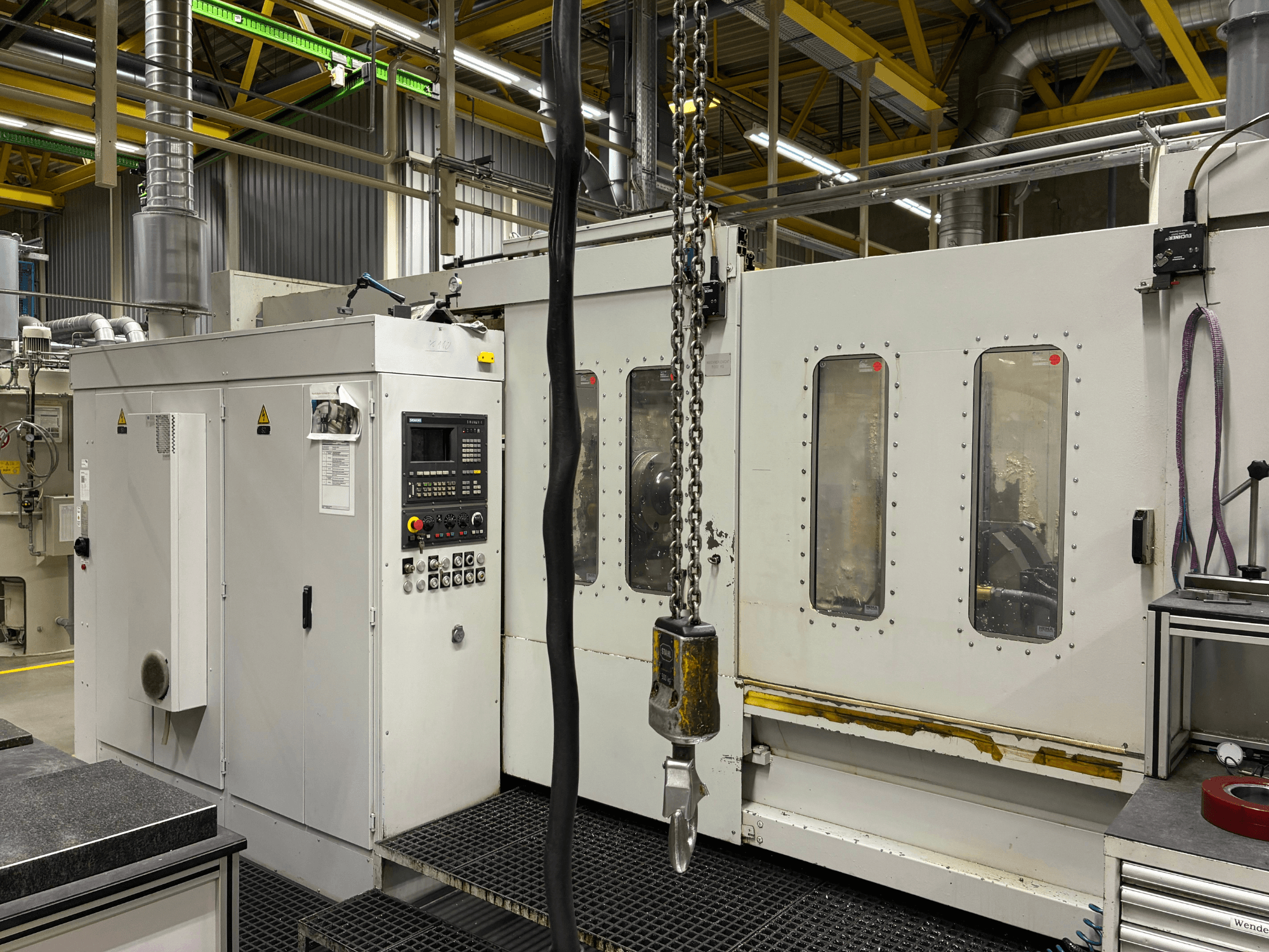 Промышленный станок с ЧПУ с панелью управления, вид сбоку; окна, цепи и металлические детали в заводских условиях.